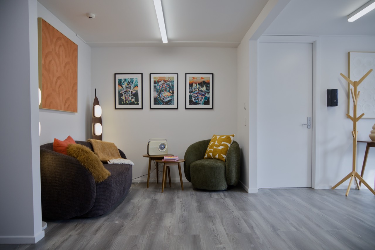 The lounge corner: a dark sofa by a tall lamp, three framed prints, a vintage radio and a green armchair
