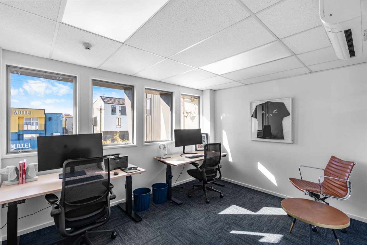 A bright studio with two desks, monitors and a row of windows
